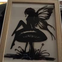 Tableau à cadre en bois clair montrant la silhouette noire d'une fée ailée assise sur un champignon, au-dessus de hautes herbes.